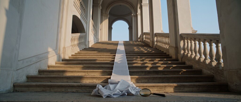 Rolo de papel impresso descendo uma escadaria de pedra, com uma lupa no último degrau.