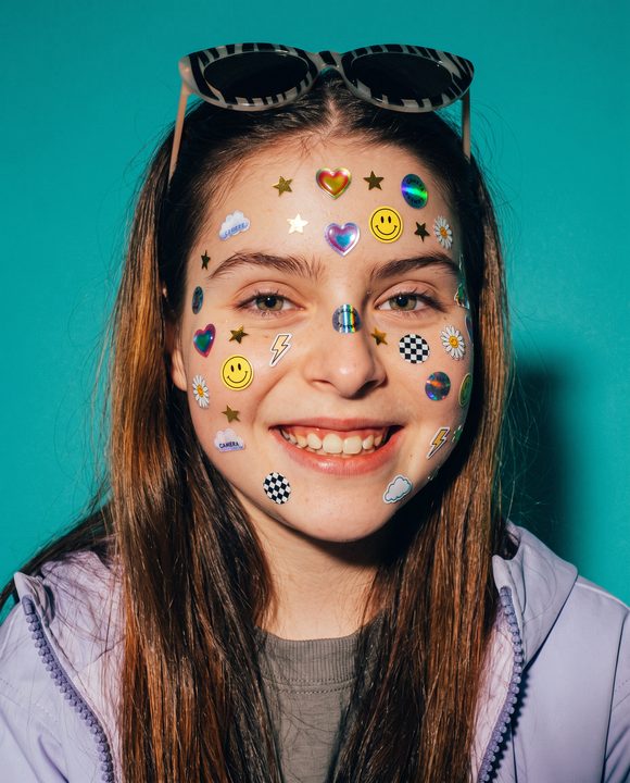 Mulher sorrindo com dezenas de adesivos coloridos colados no rosto, contra fundo verde-água