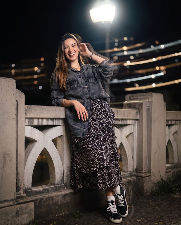 Mulher de vestido estampado encostada numa mureta à noite, com flash e luzes do fundo arrastadas