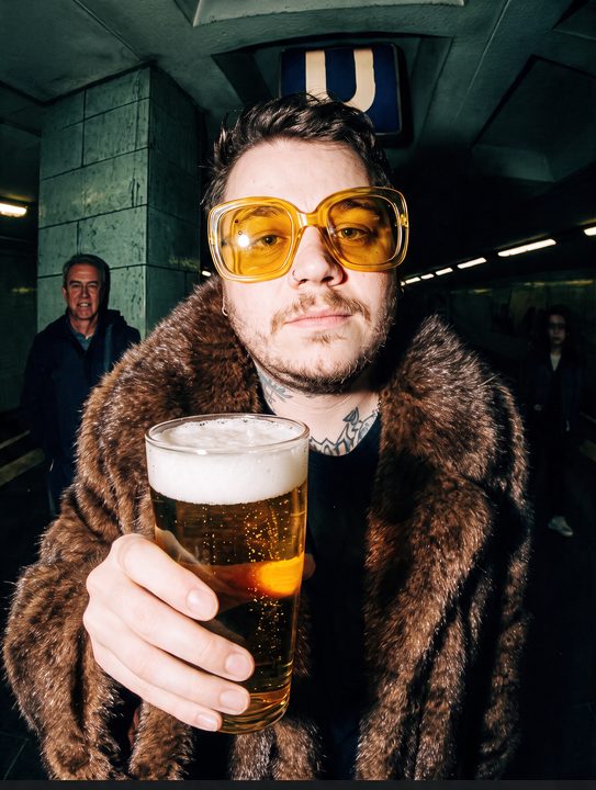 João Felipe de óculos amarelos e casaco de pele estendendo um copo de cerveja quase na lente, numa estação de metrô, com flash direto