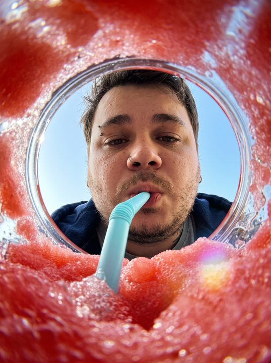 Rosto de João Felipe visto do fundo de um copo de raspadinha vermelha, com canudo azul e gelo em volta