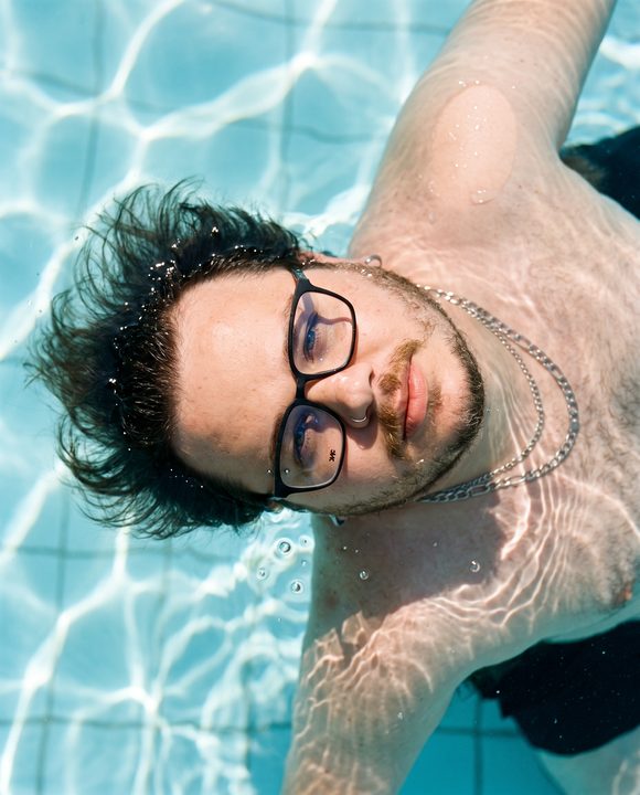 João Felipe de óculos boiando de costas na piscina, visto de cima, com reflexos do sol na água