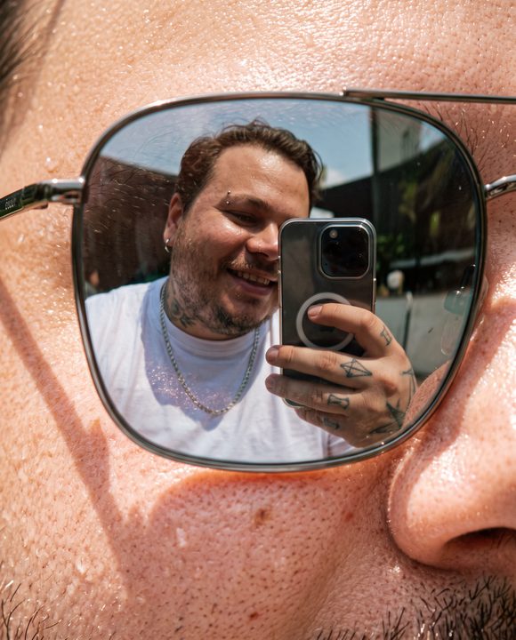 Selfie de João Felipe vista no reflexo da lente de um óculos de sol, em macro